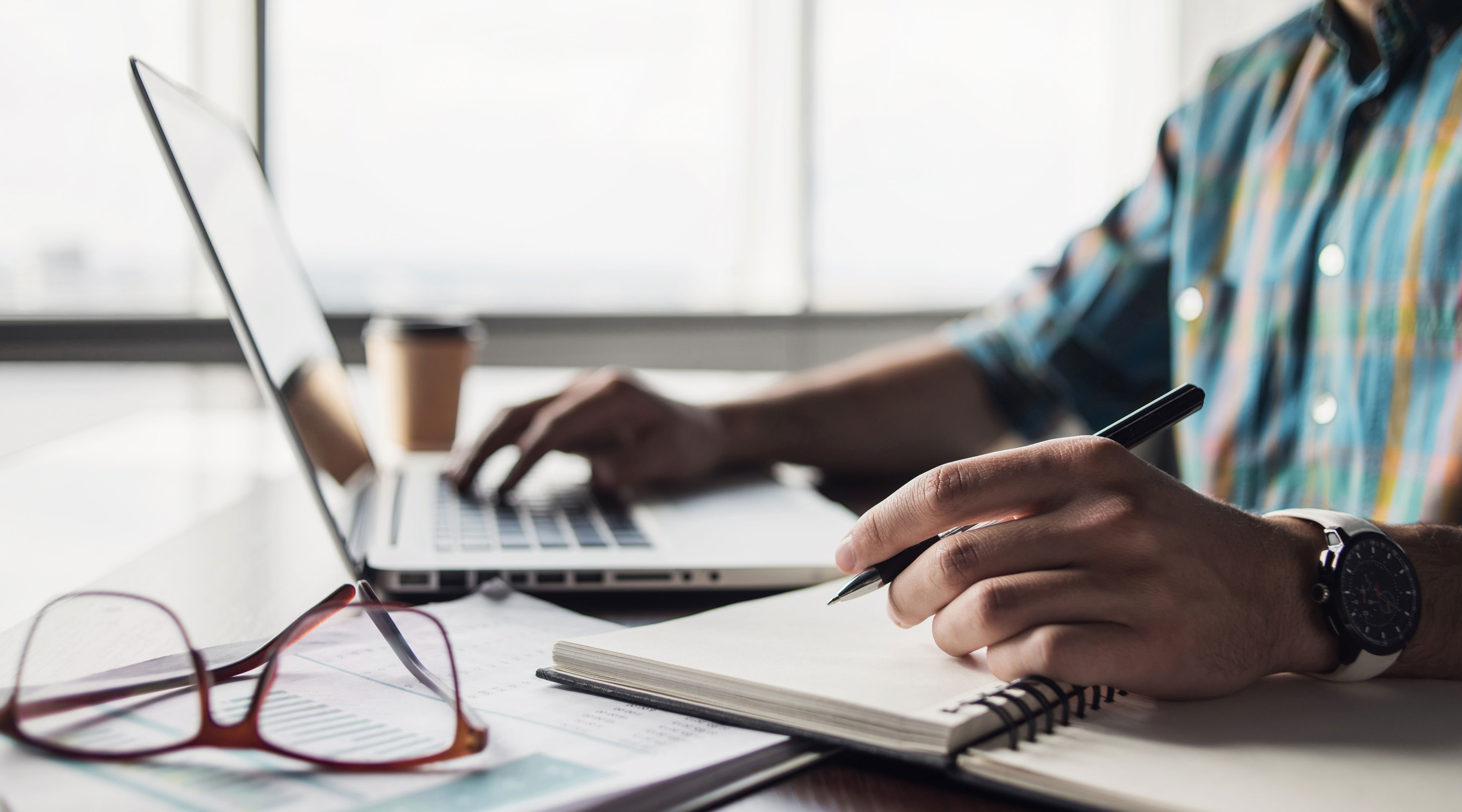 Man typing on a computer and writing on a notepad at a desk