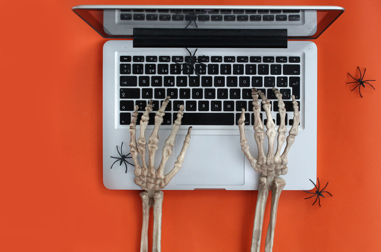 Skeleton hands typing on a laptop keyboard with small plastic spiders nearby, set against an orange background
