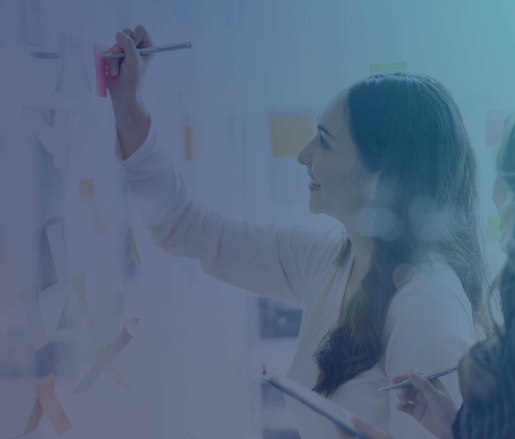 Woman in pink sweater writing on sticky notes during a collaborative planning session, smiling as she posts a red note to a glass board filled with colorful notes.
